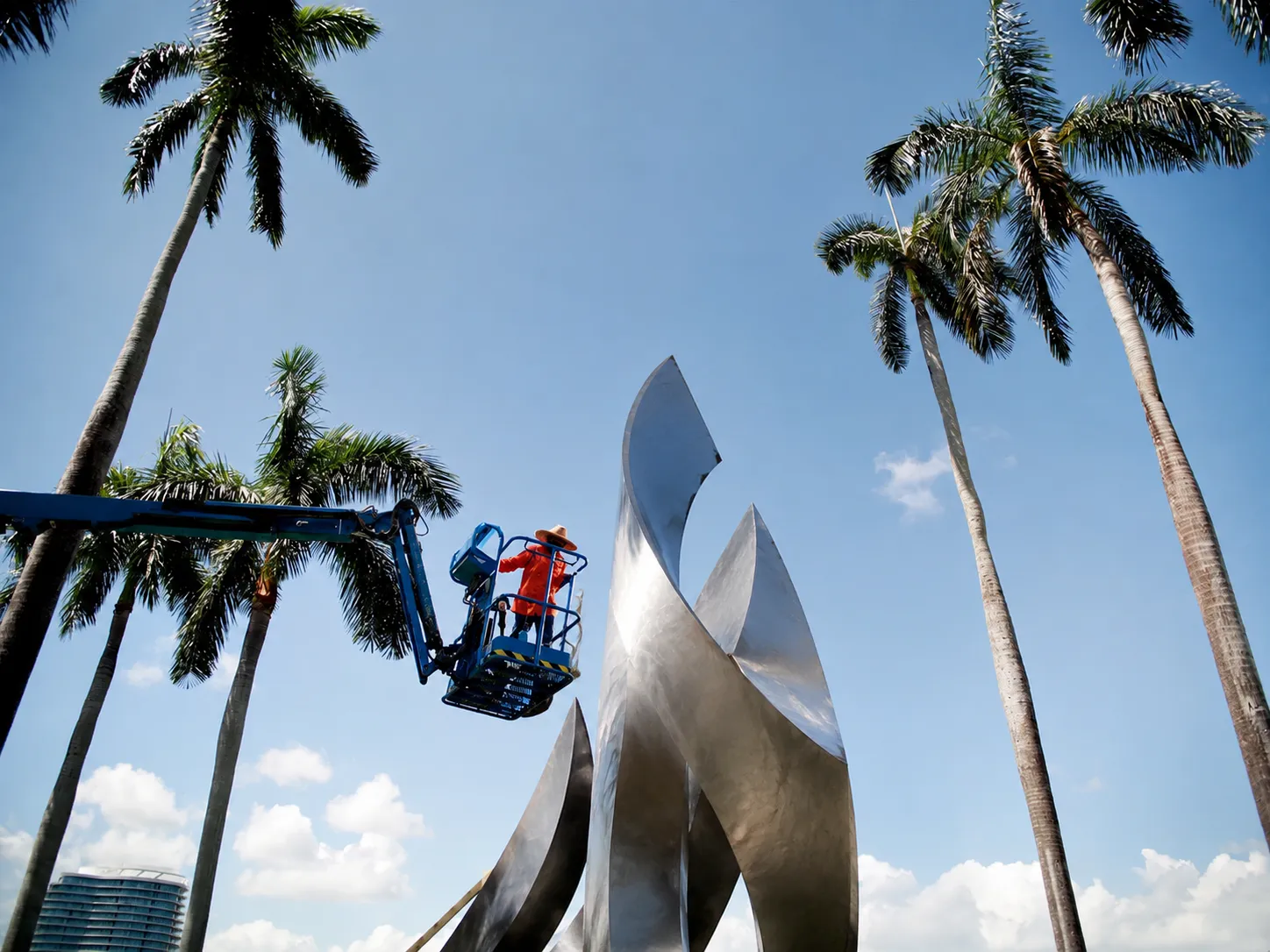 Worker maintaining a large outdoor sculpture beneath palms and open sky.